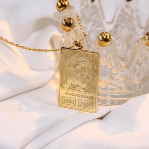 May include: A gold necklace with a pendant featuring a detailed image of a dragon with the text "3000 2500" on the bottom. The necklace is resting on a white surface.