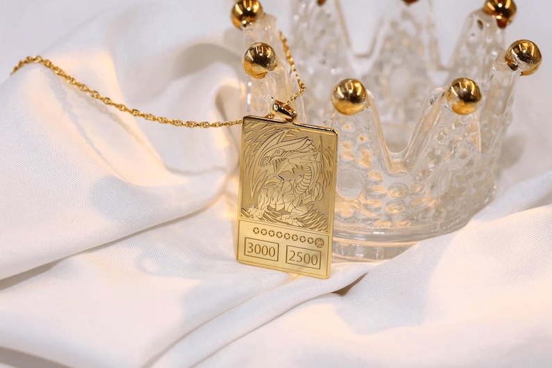 May include: A gold necklace with a pendant featuring a detailed image of a dragon with the text "3000 2500" on the bottom. The necklace is resting on a white surface.