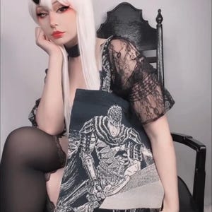 May include: A black tote bag featuring a detailed black and white illustration of a warrior. The bag is held in front of a person wearing a black lace top and black stockings. The person is seated on a black chair.