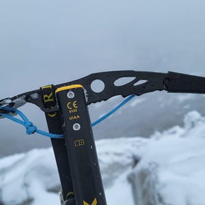 May include: Close-up of a black ice axe with a yellow accent and a blue safety cord. The ice axe is positioned against a snowy, overcast mountain backdrop. The axe has a curved pick and a handle for gripping.