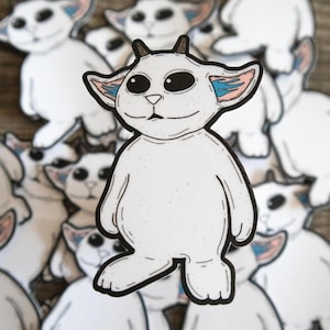 May include: White cartoon creature stickers with black outlines, large black eyes, small horns, and blue and pink accents on the ears. The stickers are scattered on a wooden surface.