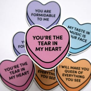 May include: A collection of heart-shaped stickers in various colors, including pink, purple, and orange. Each sticker features a black outline and a different romantic phrase, such as "You're the tear in my heart."