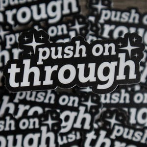 May include: Black and white stickers with the phrase "push on through" in a bold, stylized font. The stickers have a starburst design and are scattered across a wooden surface.
