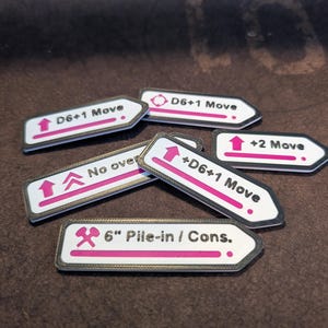 Op de afbeelding: Een set van zes witte en zwarte plastic speltokens met roze tekst. De tokens bevatten verschillende symbolen en tekst, waaronder "D6+1 Move", "No over", "6" Pile-in / Cons."