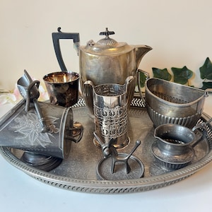 Peut inclure: Un assortiment de pièces de service en argent antiques, dont une théière, une crémière, un sucrier et un plateau de service. Les pièces sont disposées sur un plateau rond avec un bord décoratif. La théière a une poignée noire et un couvercle détaillé.
