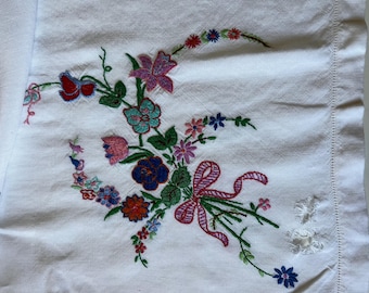 May include: A white fabric textile featuring a colorful floral embroidery pattern. The design includes various flowers in shades of red, pink, blue, purple, and green, accented by a pink ribbon tied in a bow. The fabric has a hemmed edge.