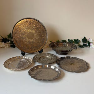 May include: A collection of antique silver serving dishes, featuring a large round tray with intricate gold patterns, smaller decorative trays, and a handled bowl. The items are displayed on a white surface.