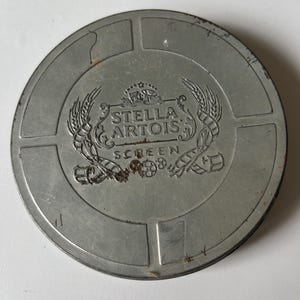 Può includere: Un contenitore rotondo in metallo color argento, con il logo e il testo Stella Artois. Il logo è centrato e presenta uno stemma con spighe di grano. Il contenitore è diviso in sezioni e ha un aspetto leggermente usurato.