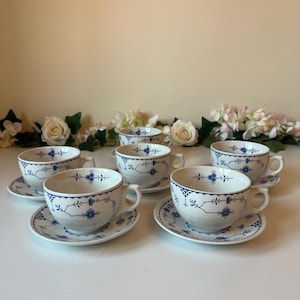 May include: A set of six white porcelain teacups and saucers. Each cup and saucer features a blue floral design. The cups have handles and sit on matching saucers. The set is arranged on a white surface.