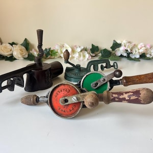 Könnte beinhalten: Eine Sammlung antiker Handwerkzeuge, darunter ein Klemmschleifer, ein grüner und schwarzer Bohrer und zwei Handbohrer mit Holzgriffen. Die Werkzeuge sind auf einer weißen Oberfläche mit Blumendekorationen angeordnet.