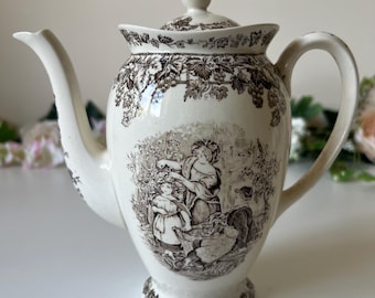 May include: A cream-colored ceramic teapot featuring a dark brown transferware print. The design includes a central scene of figures in a garden, a grape vine border around the top rim, and a matching floral pattern on the base and lid handle.