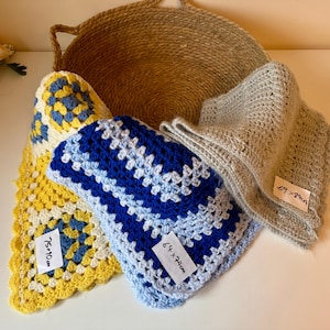 Puede incluir: Varias mantas de ganchillo hechas a mano en varios colores, incluyendo amarillo, azul y gris, cerca de una cesta tejida. Una manta está etiquetada 64 x 74cm, otra 75 x 90cm. Las mantas tienen un diseño de granny square.