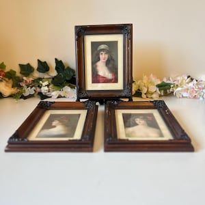 Puede incluir: Tres retratos antiguos enmarcados. Los retratos presentan mujeres con rasgos suaves y están enmarcados en madera marrón oscura con detalles ornamentados. Los retratos se exhiben sobre una superficie blanca con detalles florales.