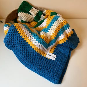 Peut inclure: Une couverture au crochet faite à la main dans un panier. La couverture présente des rayures bleues, jaunes, blanches et turquoise, avec une bordure bleue unie. La couverture mesure 205 cm x 175 cm.