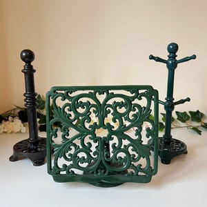 May include: Three cast iron kitchen accessories: a black paper towel holder, a green napkin holder with an ornate design, and a green mug tree. The napkin holder is square with a swirling, symmetrical pattern.