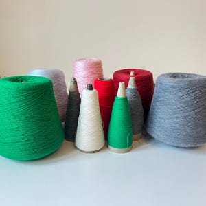 Op de afbeelding: Een verzameling kleurrijke garenkegels in verschillende tinten, waaronder groen, roze, rood en grijs. Het garen is strak op kartonnen kegels gewikkeld en toont verschillende texturen en tinten. Ideaal voor brei- en haakprojecten.
