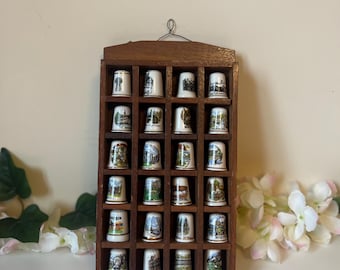 May include: A wooden display case filled with 20 decorative porcelain thimbles. Each thimble features a different design, including buildings, landscapes, and crests. The display case is rectangular with individual compartments for each thimble.