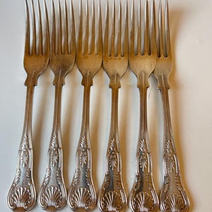 May include: A collection of seven antique silver forks. Each fork has a decorative handle with an ornate, shell-like design. The tines are long and slender. The forks are arranged in a row.