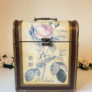 Könnte beinhalten: Eine Vintage-Holzkiste mit Scharnierdeckel, mit einem Blumendesign einer rosa Rose und botanischen Illustrationen. Die Kiste hat einen dunkelbraunen Rahmen, goldfarbene Verzierungen sowie einen Metallgriff und -verschluss. Text: "Rosier mousseux" und "à fleurs doubles".