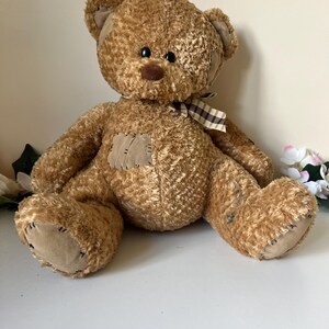 Può includere: Un orsacchiotto marrone con un corpo strutturato, toppe e un nastro a quadri. L'orso ha un naso marrone, occhi neri e zampe color cuoio. L'orso è seduto su una superficie bianca.