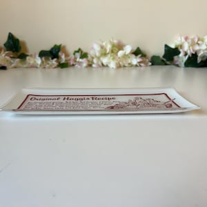 May include: Rectangular glass serving dish with a white background and a maroon border. The dish features the text "Original Haggis Recipe" in maroon, along with a detailed illustration. Decorative flowers are in the background.