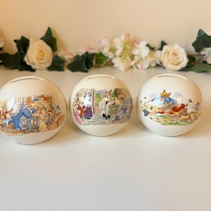 Può includere: Tre salvadanai rotondi in ceramica color crema con illustrazioni di scene di Peter Coniglio. Ogni salvadanaio ha una fessura sulla parte superiore per inserire le monete. Composizioni floreali di fiori bianchi e rosa sullo sfondo.