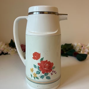 May include: A vintage thermos flask in white and pale green, with a handle and spout. It has a floral design of red roses and yellow flowers. The top and handle are white, and the body is pale green with a decorative stripe.