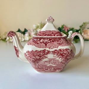 Può includere: Teiera vintage con base color crema e design rosso. La teiera presenta una scena paesaggistica dettagliata e motivi floreali. Ha un beccuccio curvo, un manico e un coperchio con un pomello.