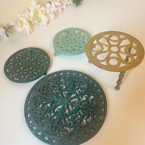 May include: A collection of metal trivets in different shapes and colours. There's a round, dark green trivet with an intricate pattern, a light green trivet, and a gold-coloured trivet with a floral design. A sprig of flowers is in the background.