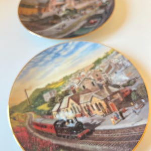 May include: Two decorative plates with gold rims. The plates depict a detailed landscape scene with a train, buildings, and a cloudy sky. The plates are round and appear to be made of porcelain.