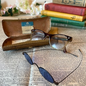 May include: A pair of vintage spectacles with brown frames and clear lenses sits on a newspaper. Another pair with blue lenses is below. An open brown case with a Mopar label and a stack of old books are visible.