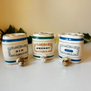 May include: Three ceramic barrel-shaped liquor dispensers. Each is white with blue and gold bands, and a brass spigot. One reads "GIN", another "SHERRY", and the third "WHISKY". Decorative and vintage-inspired.