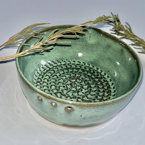 May include: A round, light green ceramic grater bowl with a textured, dark brown spiral pattern at the base for grating. The bowl features a smooth, glossy glaze, a thick rim, and three small circular indentations along the exterior side wall.