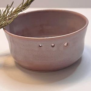 May include: A small, round, dusty rose-colored ceramic bowl with three evenly spaced holes near the top rim. A sprig of dried greenery rests near the bowl. The bowl has a smooth, glossy finish and a slightly flared shape.