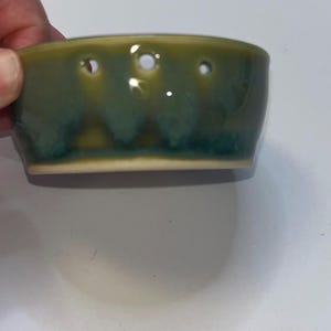 May include: A small, round ceramic bowl featuring a glossy glaze in shades of olive green and deep teal. The upper rim of the bowl is punctuated by a series of evenly spaced, small circular holes that encircle the upper portion of the vessel.