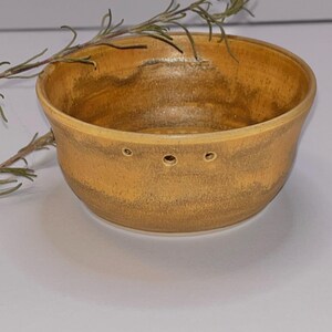 May include: A handmade, ceramic bowl with a warm, earthy color palette. The bowl features a rounded shape with a slightly flared rim and three small holes near the base. A sprig of rosemary rests on the rim, adding a natural touch.