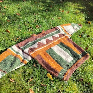 Op de afbeelding: Een kleurrijke trui met patronen in groen, bruin, oranje en wit, liggend op groen gras. De trui heeft geometrische ontwerpen en een geribbelde halslijn en manchetten. De trui is gemaakt van dik, getextureerd garen.