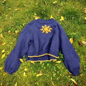 Op de afbeelding: Een marineblauwe gebreide trui met een gele zon op de rug. De trui heeft lange mouwen en een cropped pasvorm, met gele rand. Hij ligt op groen gras met gevallen bladeren. Een comfortabel kledingstuk.