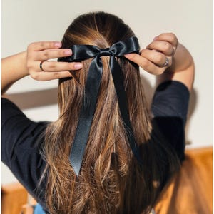 May include: A black satin hair bow with long streamers is tied in the hair. The bow is centered at the back of the head, with the streamers cascading down the hair. The hair is long, wavy, and a mix of brown and blonde.