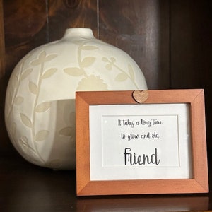 May include: A wooden picture frame with a white background and the text "It takes a long time to grow old friend". The frame is sitting on a wooden shelf with a white ceramic vase with floral designs behind it.