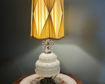 Può includere: Una lampada da tavolo in vetro bianco scanalato con una base dorata e un paralume plissettato a righe. Il paralume è di colore giallo chiaro con strisce marroni.