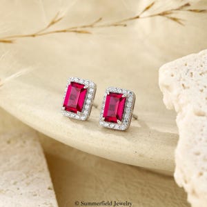 May include: A pair of stud earrings featuring rectangular, emerald-cut vibrant red center stones. Each red stone is surrounded by a halo of small, clear, round-cut white stones set in a polished silver-toned metal frame with post backings.
