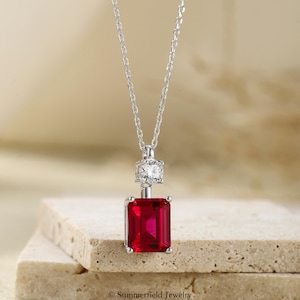 May include: A silver-toned necklace featuring a rectangular, emerald-cut red gemstone pendant suspended from a delicate chain. Above the red stone, a smaller, round, clear faceted stone is set in a silver-toned mount. The text Summerfield Jewelry is visible.