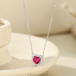 Könnte beinhalten: Eine silberne Halskette mit einem herzförmigen Anhänger, der einen zentralen roten Edelstein besitzt, welcher von einem Kranz aus kleinen klaren Steinen umgeben ist. Der Text Summerfield Jewelry ist unten im Bild in kleiner schwarzer Schrift gedruckt.