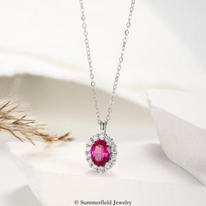 May include: A silver-toned necklace featuring a delicate chain and an oval-shaped pendant. The pendant showcases a vibrant, deep pink oval gemstone at its center, surrounded by a halo of small, clear, sparkling stones. The text Summerfield Jewelry is visible.