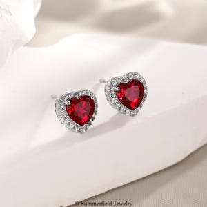 May include: A pair of heart-shaped stud earrings featuring a vibrant red center stone surrounded by a halo of small, clear, sparkling stones set in a polished silver-toned metal. The earrings are designed with a post back for secure attachment.