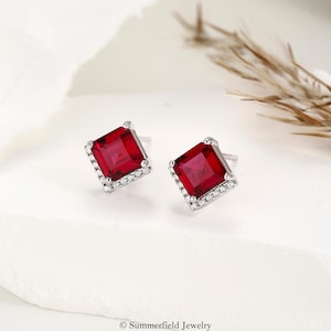 May include: A pair of square-cut red gemstone stud earrings, each featuring a central deep red stone surrounded by a halo of small clear accent stones set in a silver-toned metal frame. The text Summerfield Jewelry is visible at the bottom.