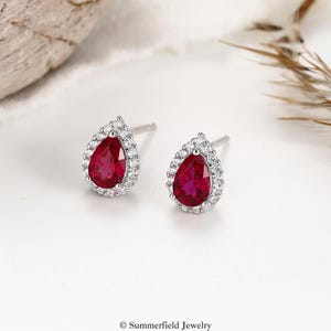 May include: A pair of pear-shaped stud earrings. Each earring features a vibrant red gemstone surrounded by a halo of small, sparkling white stones. The earrings are set on silver posts, creating a classic and elegant look.