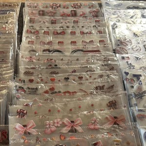 May include: A display of various sticker sheets in clear plastic packaging. The stickers feature baseball team logos, bows, hearts, and other decorative elements. The stickers are arranged in rows and columns, showcasing a variety of designs.