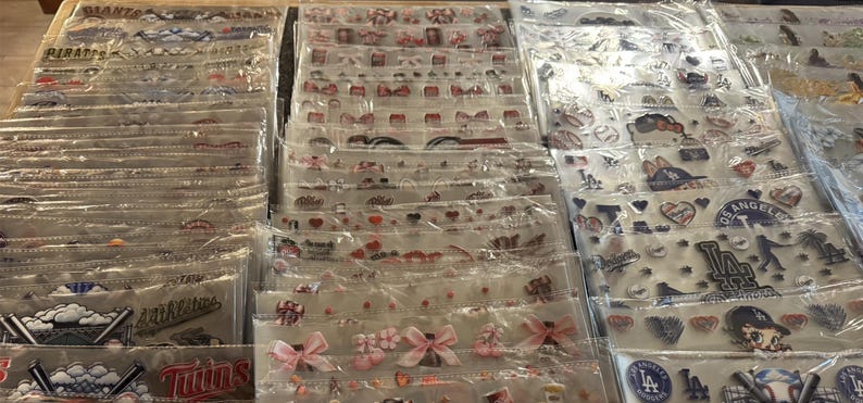 May include: A display of various sticker sheets in clear plastic packaging. The stickers feature baseball team logos, bows, hearts, and other decorative elements. The stickers are arranged in rows and columns, showcasing a variety of designs.